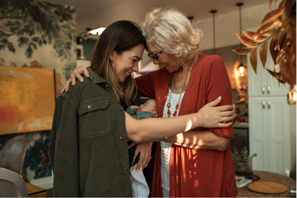 A smiling woman of European descent sits in a comfortable chair and nurses her infant while speaking with a woman of African descent.