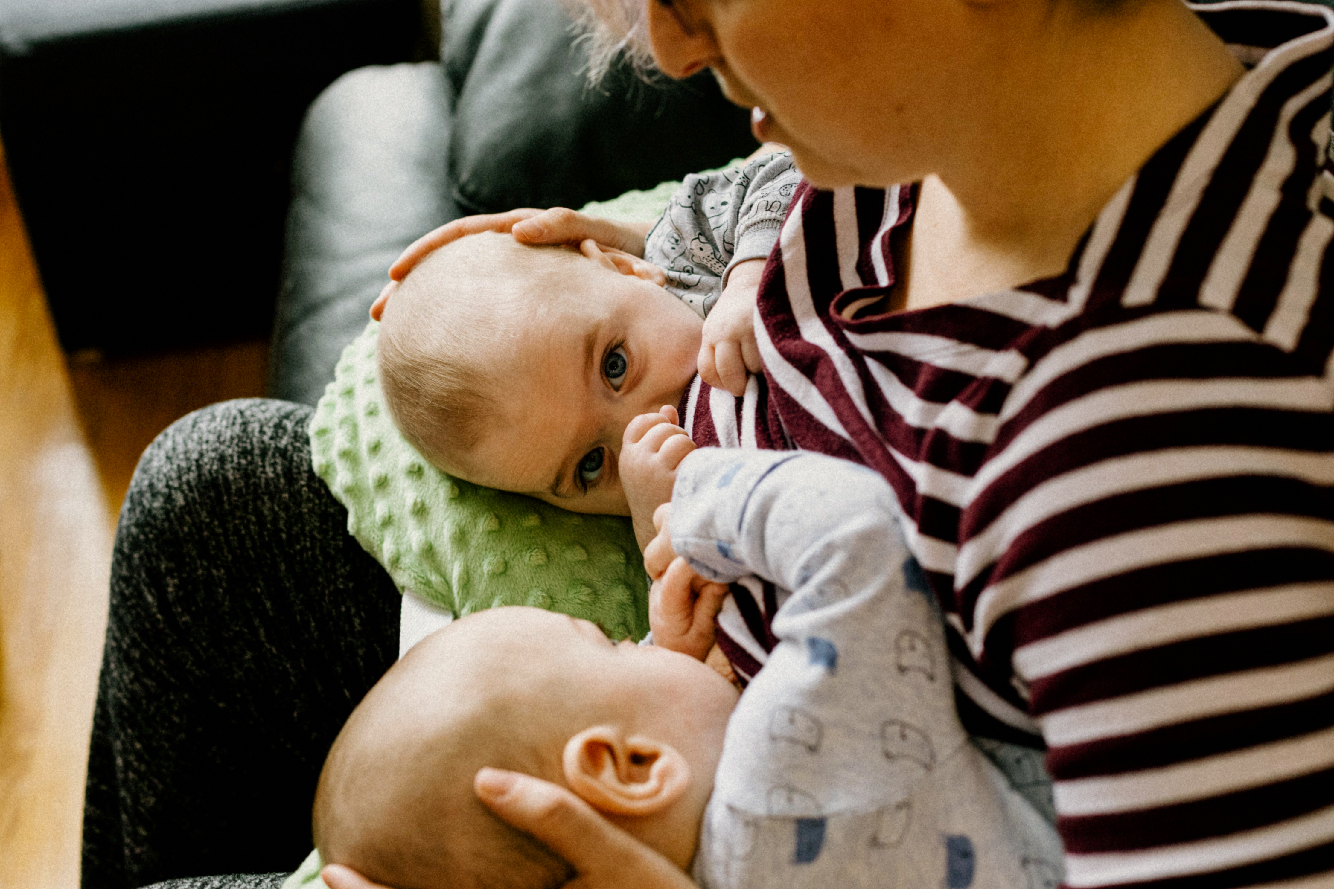 A smiling woman of European descent sits in a comfortable chair and nurses her infant while speaking with a woman of African descent.
