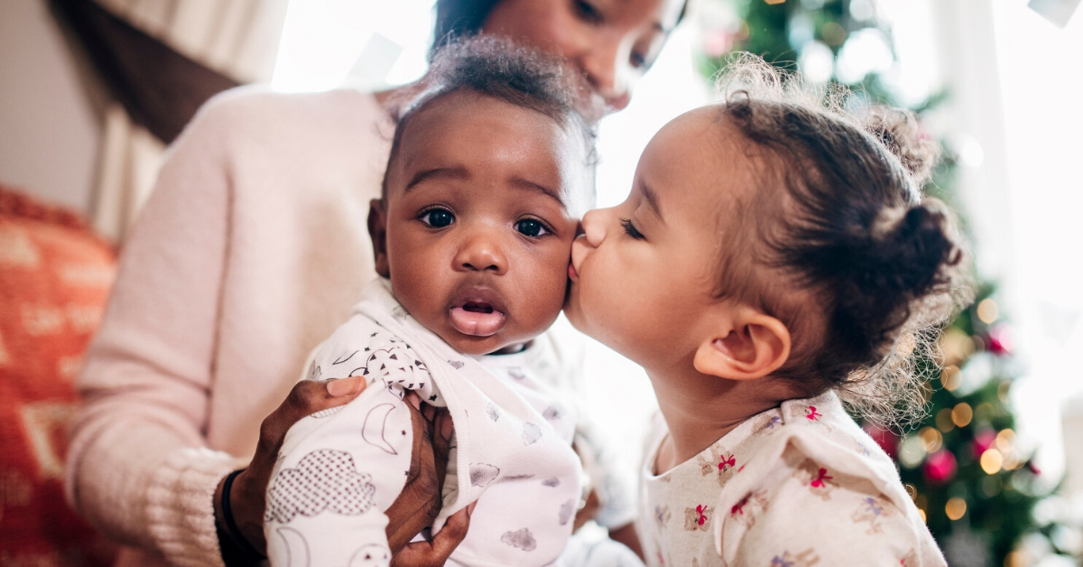 A smiling woman of European descent sits in a comfortable chair and nurses her infant while speaking with a woman of African descent.