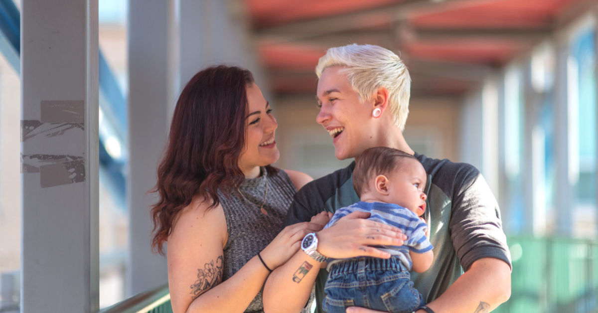 A smiling woman of European descent sits in a comfortable chair and nurses her infant while speaking with a woman of African descent.