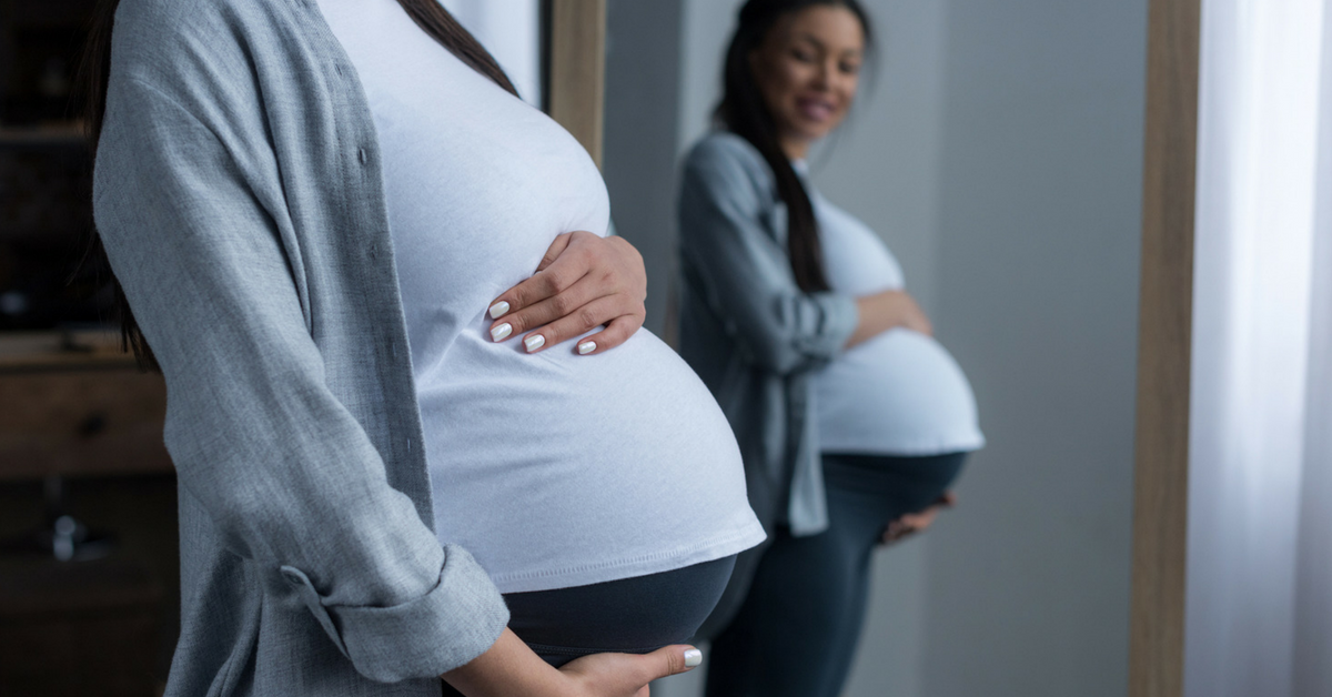 A smiling woman of European descent sits in a comfortable chair and nurses her infant while speaking with a woman of African descent.
