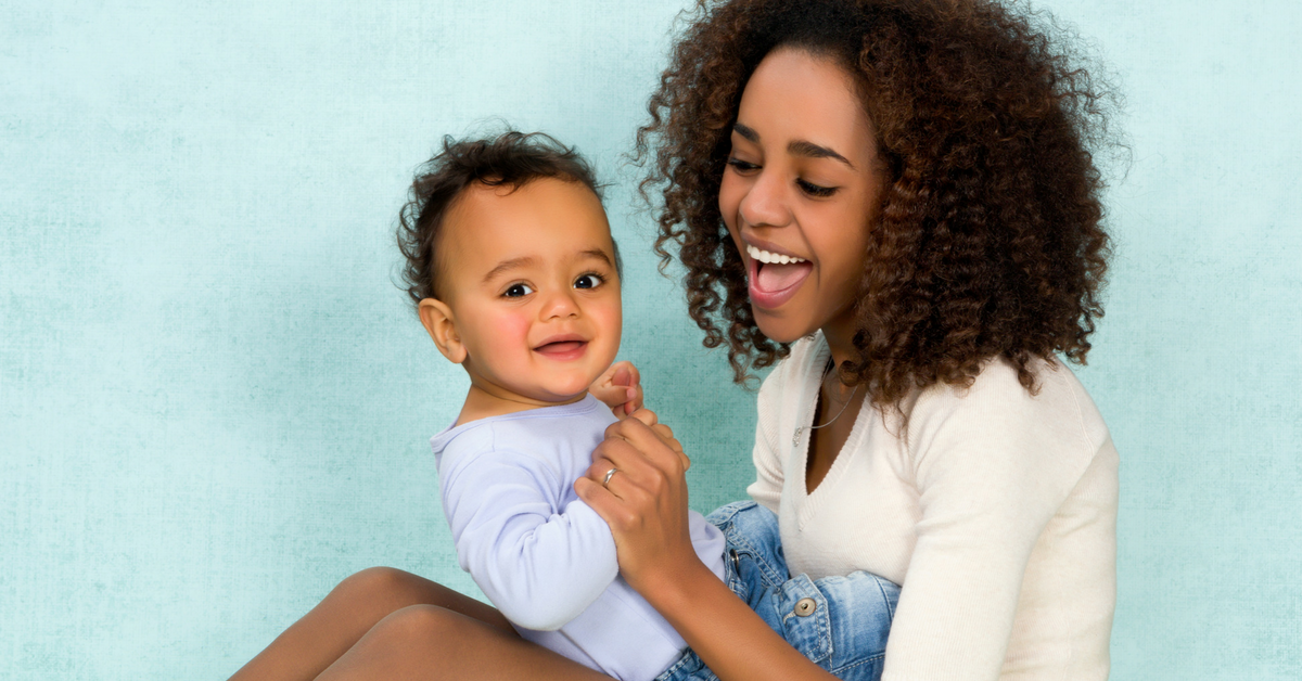 A smiling woman of European descent sits in a comfortable chair and nurses her infant while speaking with a woman of African descent.