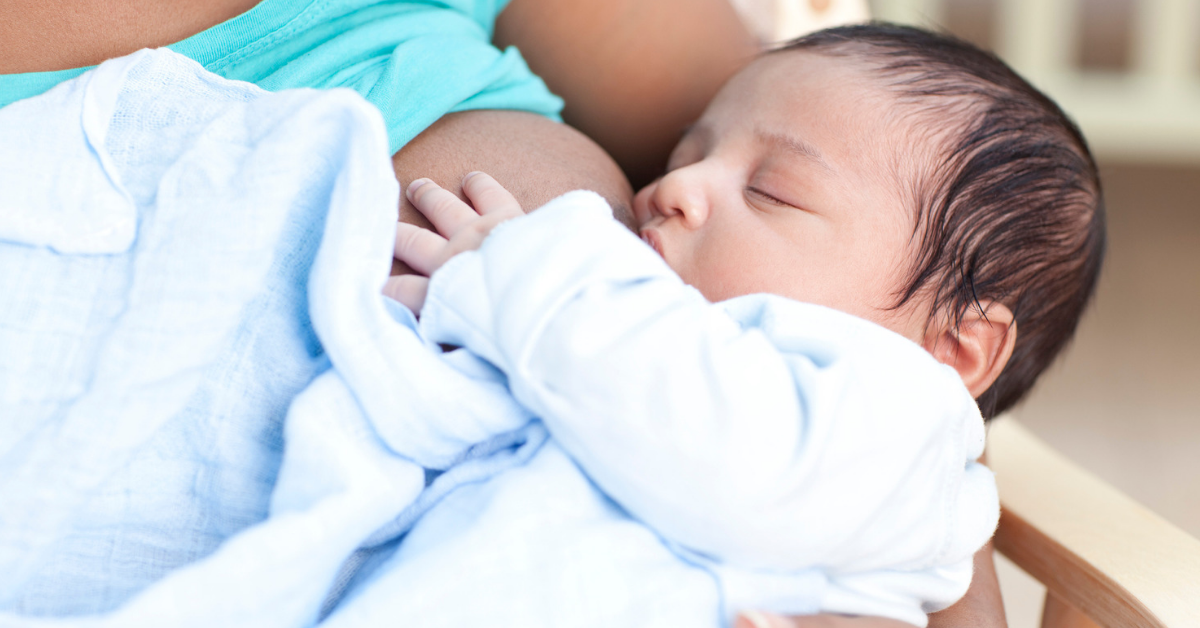 A smiling woman of European descent sits in a comfortable chair and nurses her infant while speaking with a woman of African descent.