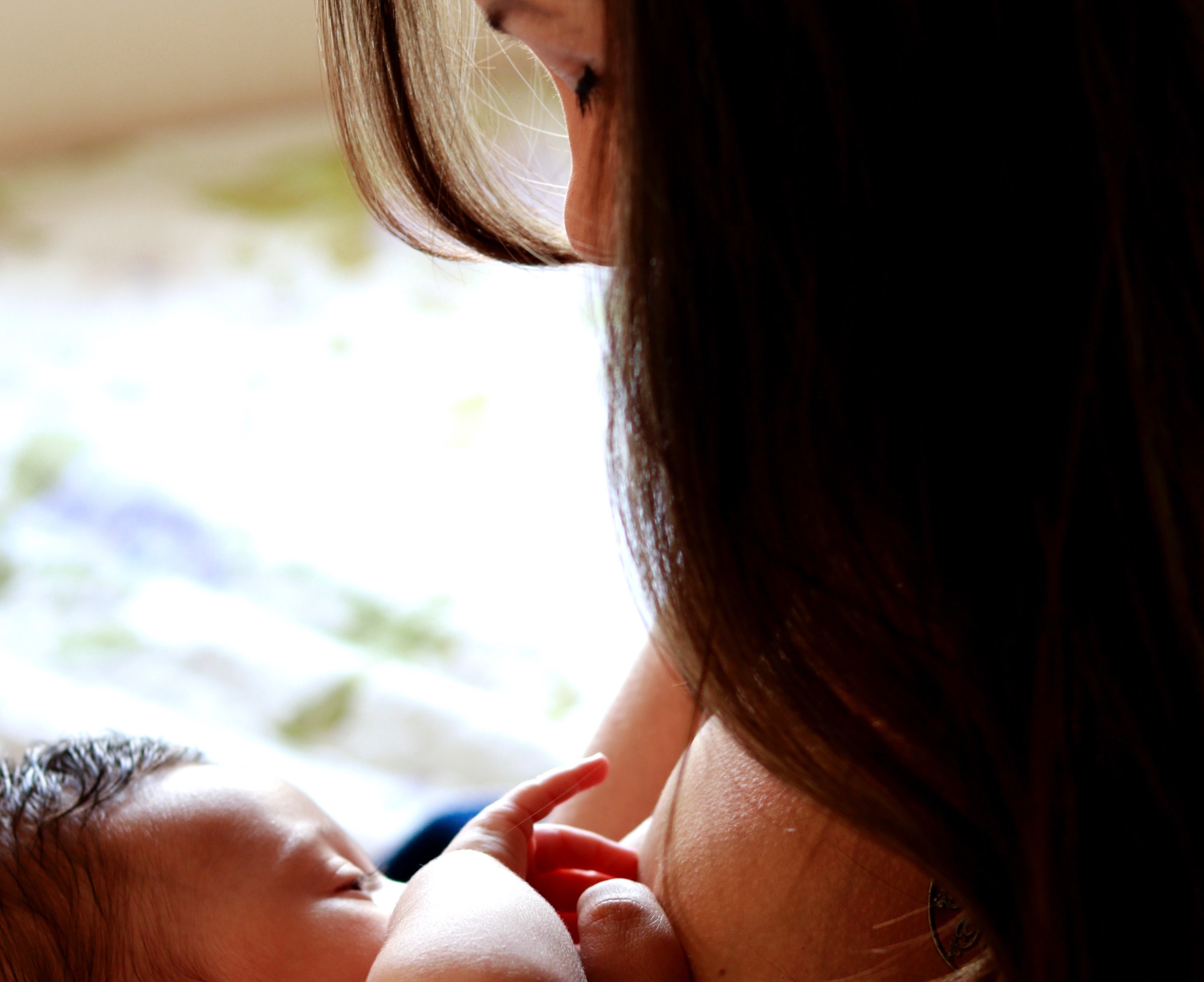 A smiling woman of European descent sits in a comfortable chair and nurses her infant while speaking with a woman of African descent.