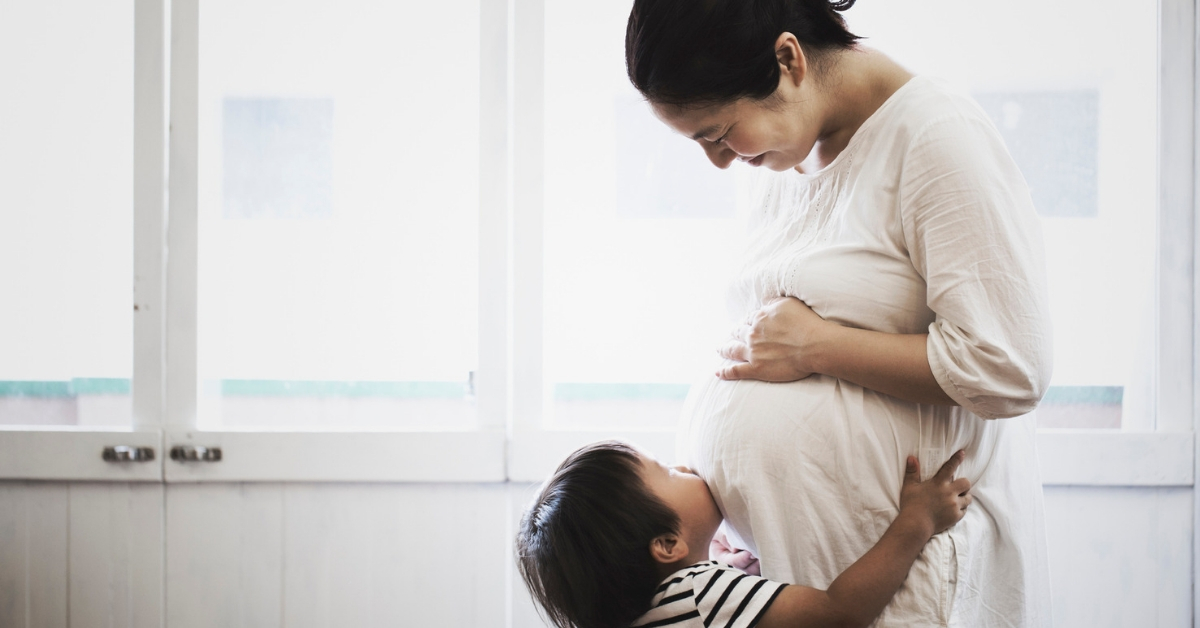A smiling woman of European descent sits in a comfortable chair and nurses her infant while speaking with a woman of African descent.