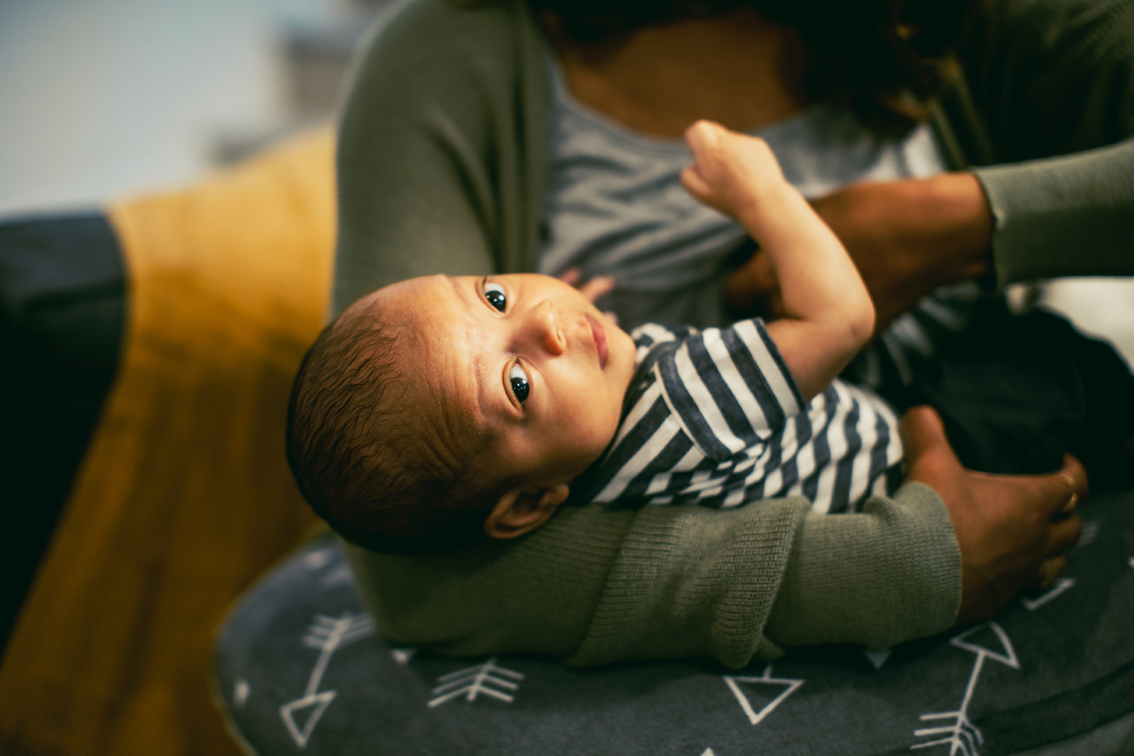 A smiling woman of European descent sits in a comfortable chair and nurses her infant while speaking with a woman of African descent.