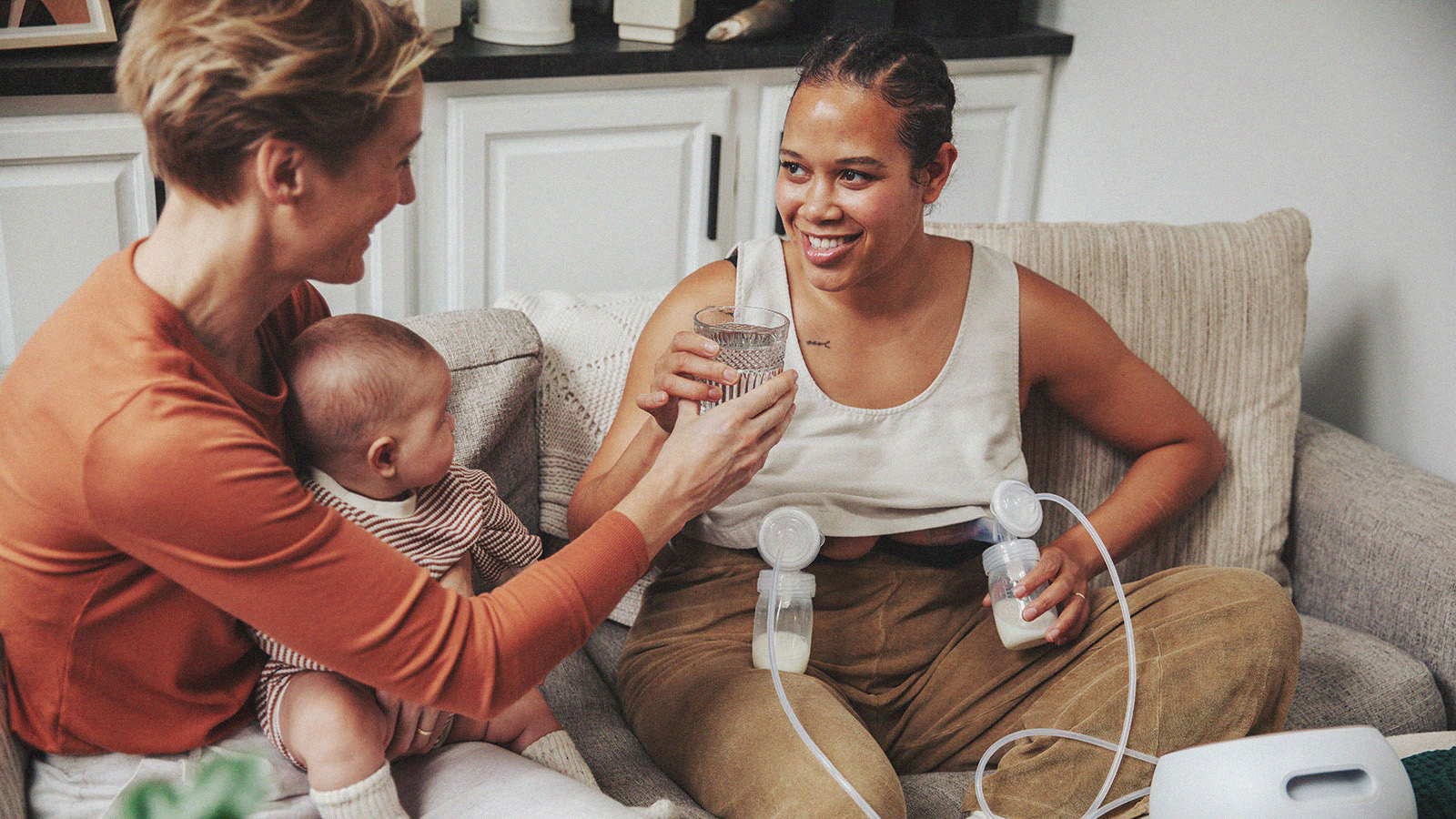 A smiling woman of European descent sits in a comfortable chair and nurses her infant while speaking with a woman of African descent.